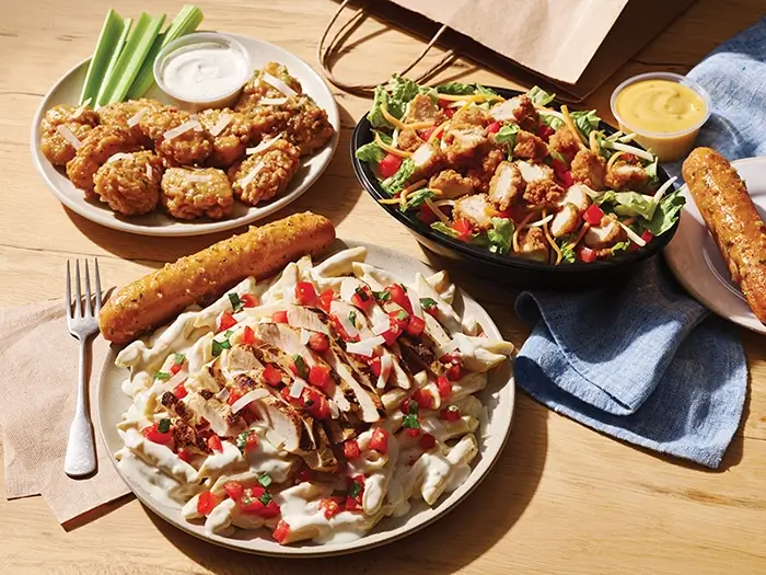 Boneless wings, chicken pasta, salad and breadsticks served with dipping sauces on a restaurant table.