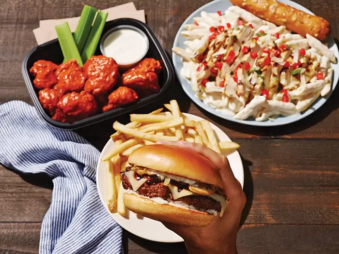 To-go wings with celery, a burger, fries and chicken pasta arranged on a restaurant table.